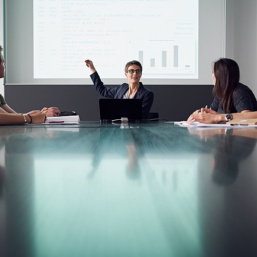 Personengruppe sitzt an einem Tisch während eine an die Wand mit dem Projektor zeigt