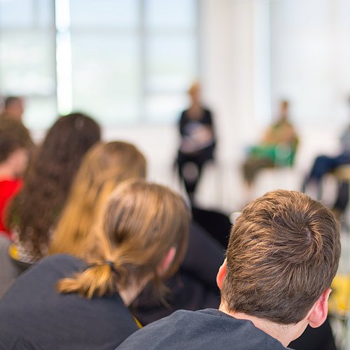 Personen hören einer Diskussion zu und im Hintergrund sitzen Personen dem Publikum zugewandt