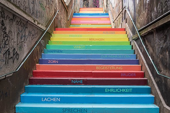 Das Bild zeigt eine Treppe mit farbigen Stufen und verschiedenen Begriffen wie Ehrlichkeit, Nähe, Sprechen uvm.